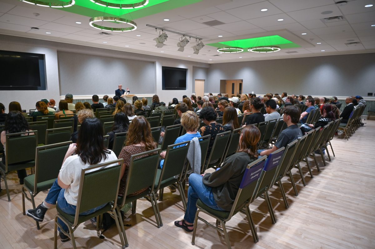 People sit in green chairs in a lecture hall with a speaker at a podium at the front.