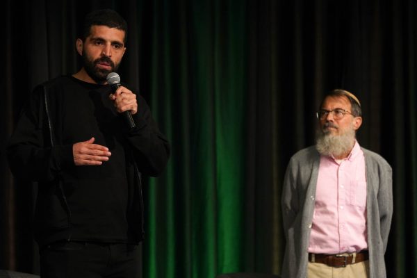 Two men stand on stage. One is wearing black clothing, speaking into a microphone, and the other is wearing a grey sweater and pink shirt. They are both standing behind a dark green curtain.