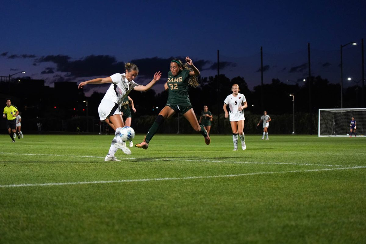 A woman in a green jersey jumps after a soccer ball while another woman in a white jersey kicks the ball.