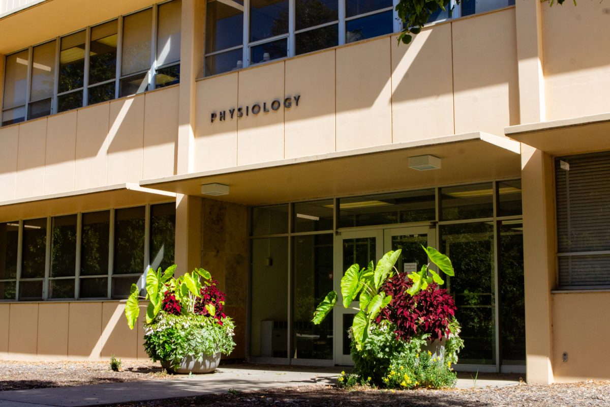 On a sunny day, a tan building with the word "Physiology" is devoid of people. There are two large plants on either side of the entry door.