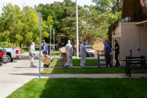 On a sunny day, people wearing protective suits and masks move boxes into the a tan building.