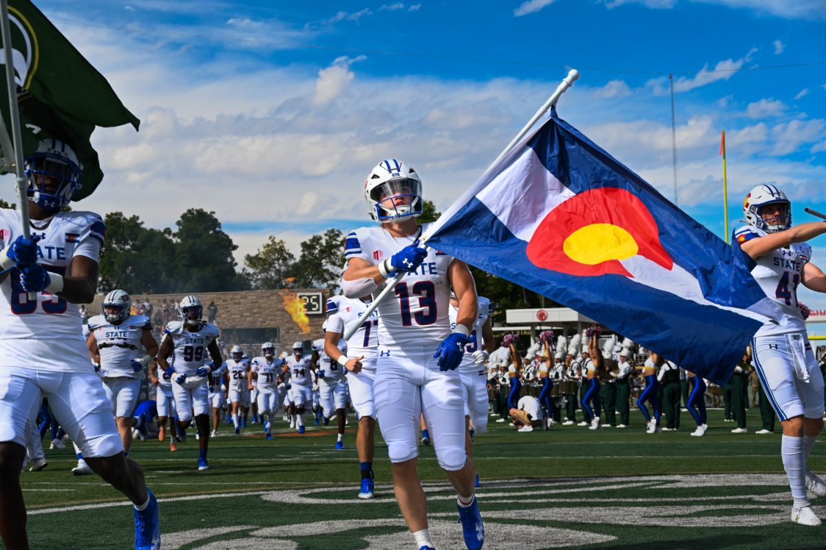 a football player in royal blue and white runs onto the field while holding the Colorado state flag