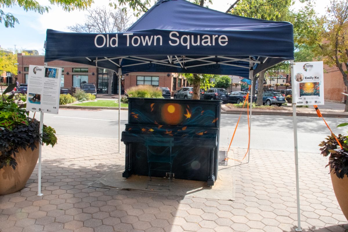 A piano that is being in the middle of being painted sits under a tent in front of a street.