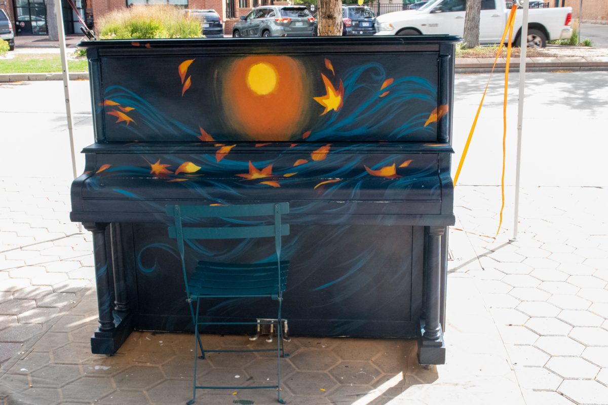 A piano that is being in the middle of being painted sits under a tent in front of a street.