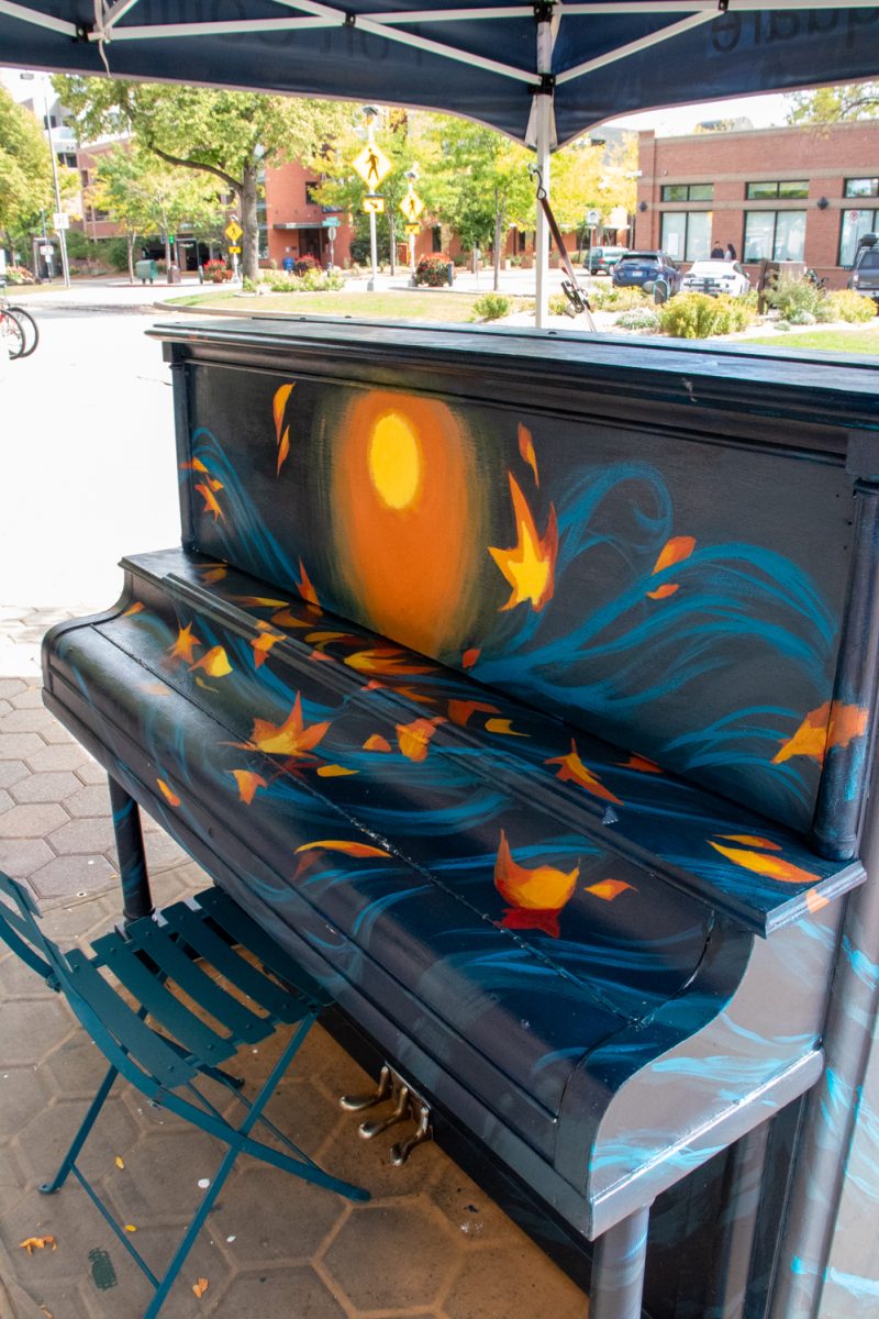 A piano that is being in the middle of being painted sits under a tent in front of a street.