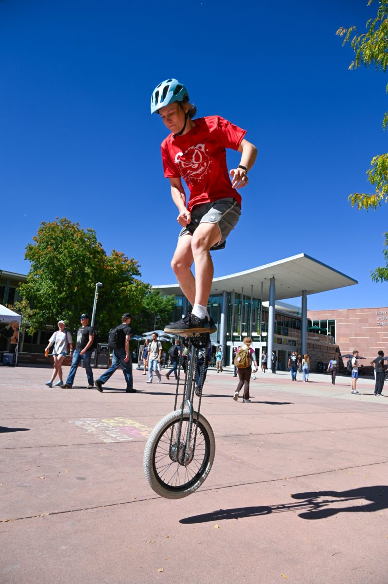 A man in a red shirt and grey shorts rides on a tall silver unicycle