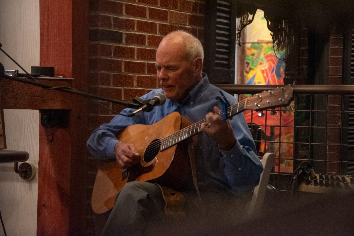 Man in blue shirt sits down and plays guitar with microphone in front of him