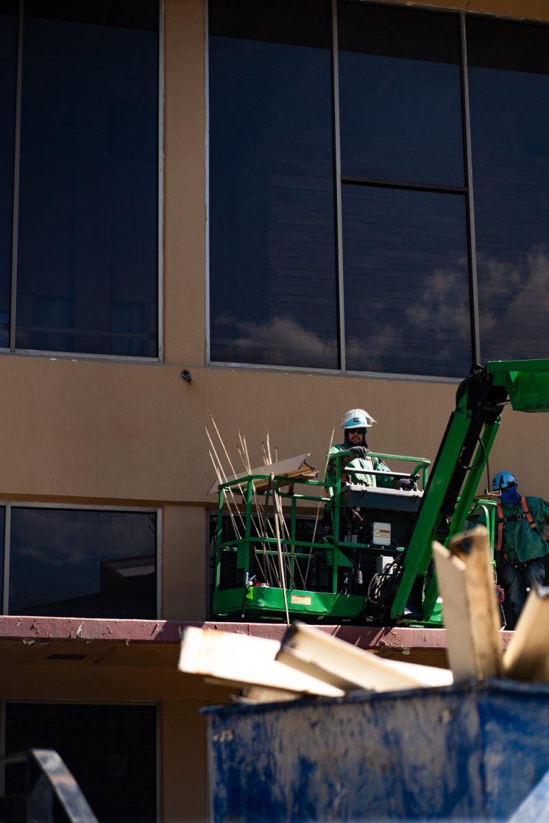 A construction worker in personal protective equipment stands on a green construction lift in front of a building.
