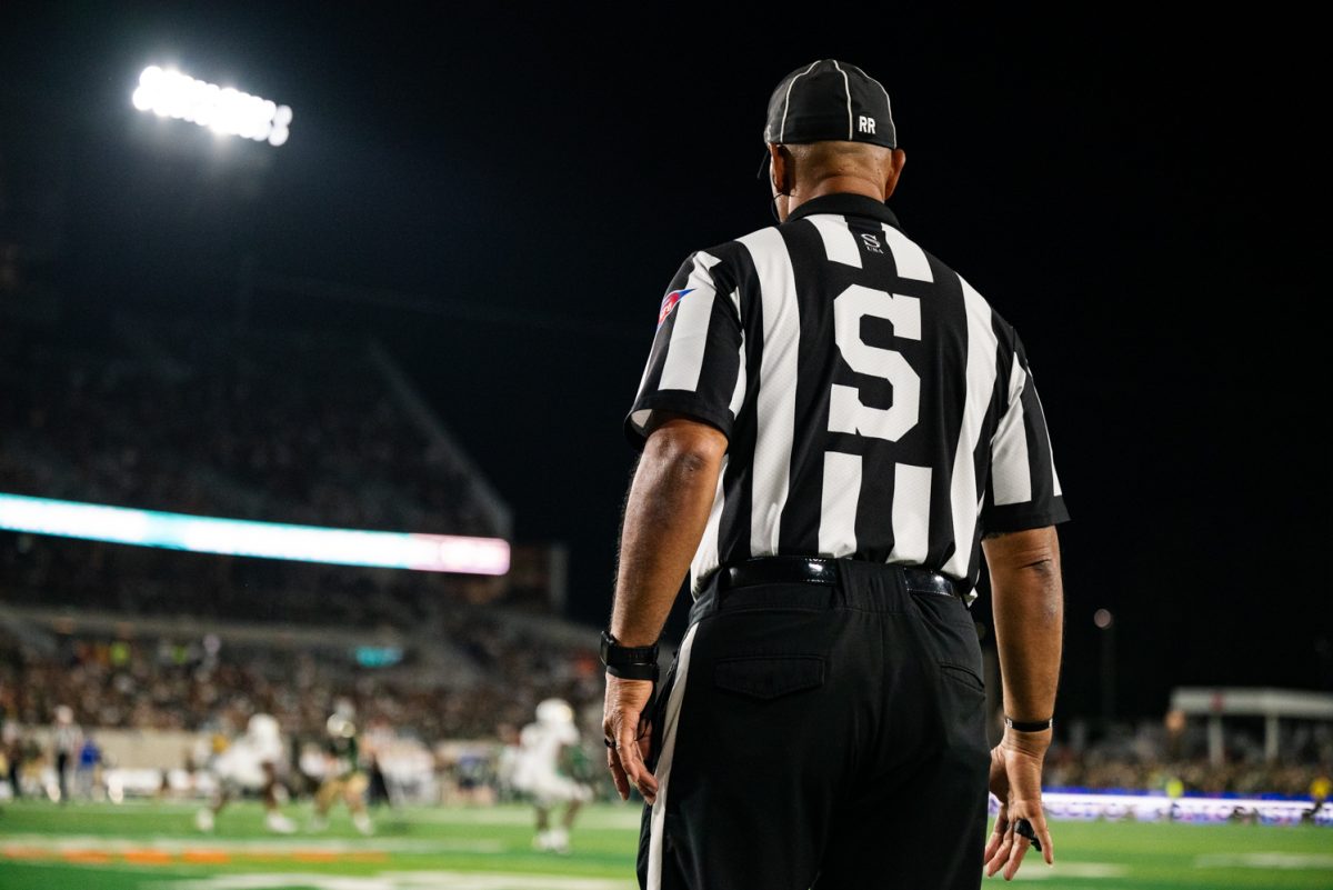 A referee in black and white stripes with an S on his back faces the football field.