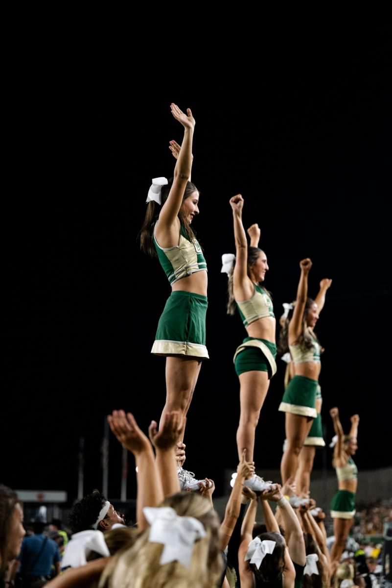 Four cheerleaders in green and gold stand on top of their stacks and wave with both arms.