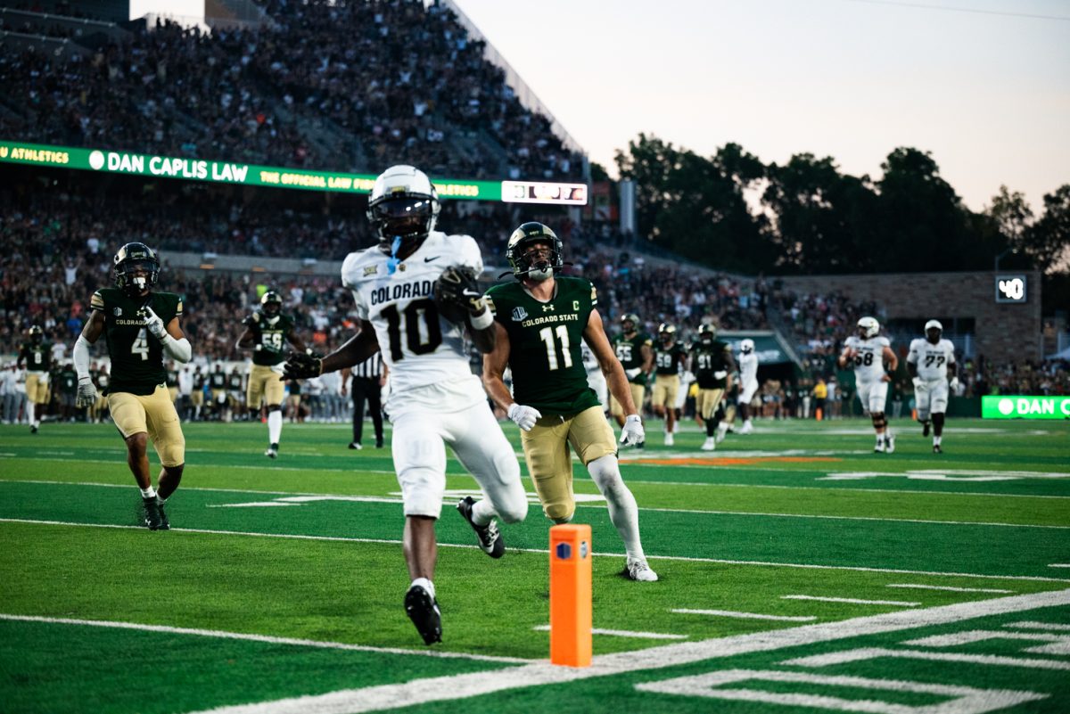 A University of Colorado player in a white and black jersey runs with the football into the end zone. He is followed by a Colorado State football player in a green and gold jersey.
