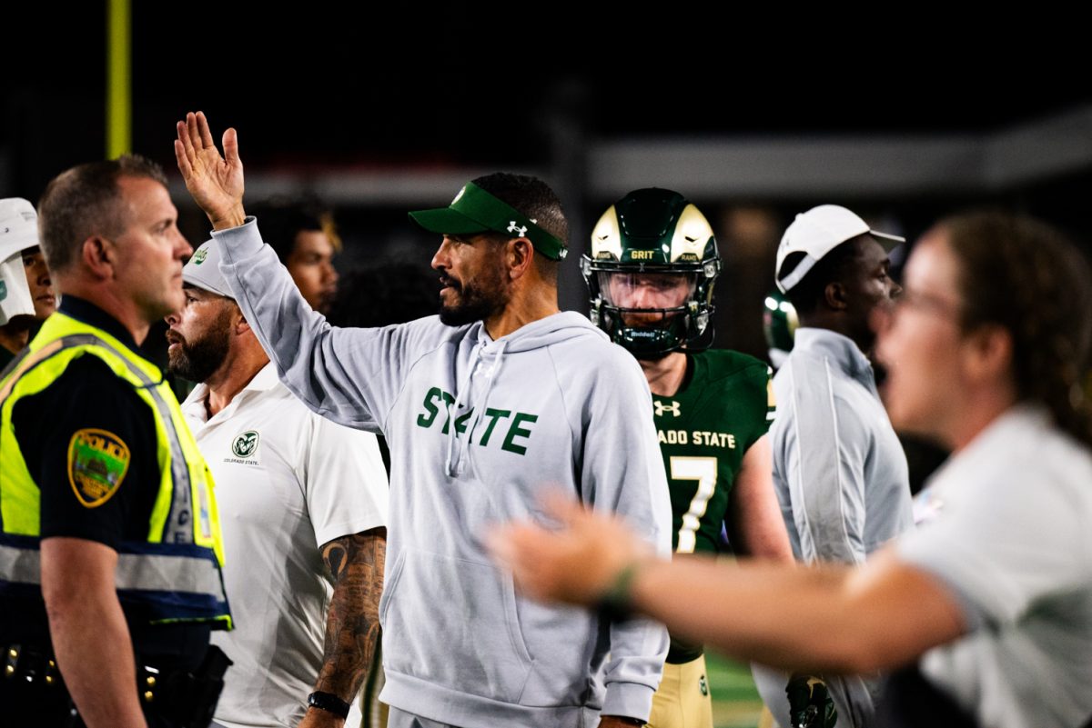 A man in a gray sweatshirt, standing among other people milling about, holds his arm up to call over his team.