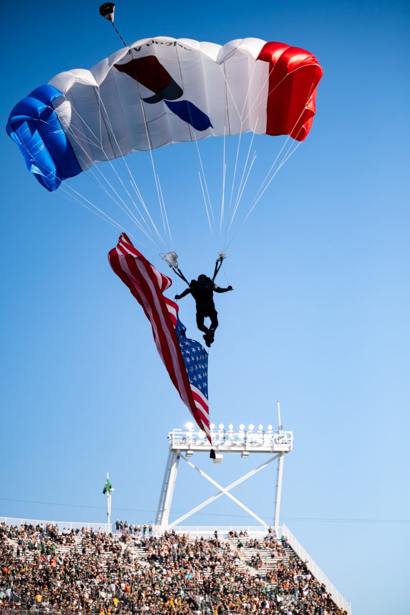 A red, white and blue paraglider carrying an American flag floats to the ground in front of a stadium of people.