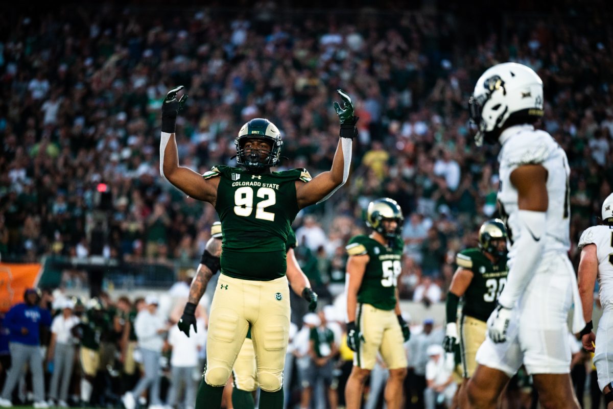 A football player in green and gold holds up his hands in front of a sea of CSU fans while a University of Boulder walks next to him.