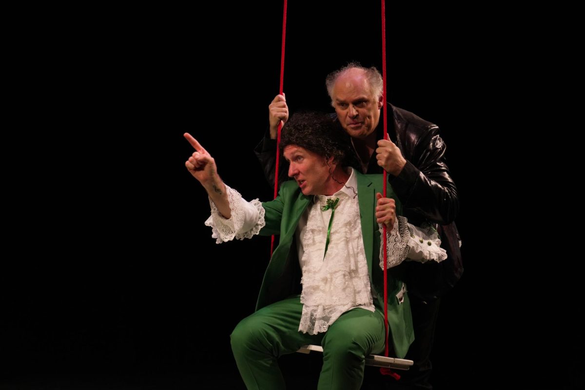 A man in a white shirt and green suit is pushed on a swing by a man in a black leather jacket