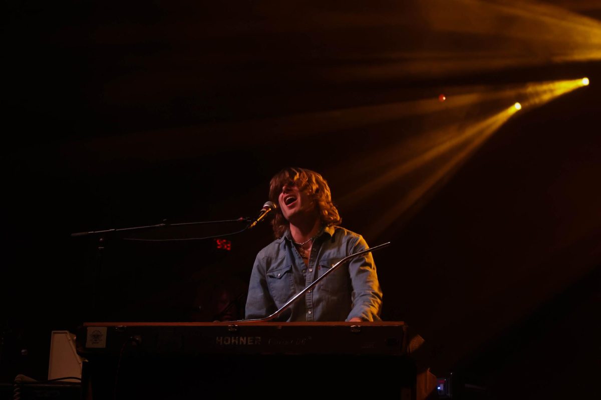 A man with shaggy brown hair sings into a microphone while playing a piano. Yellow-gold lights illuminate him from the top right corner, most of the stage is in dramatic shadow.