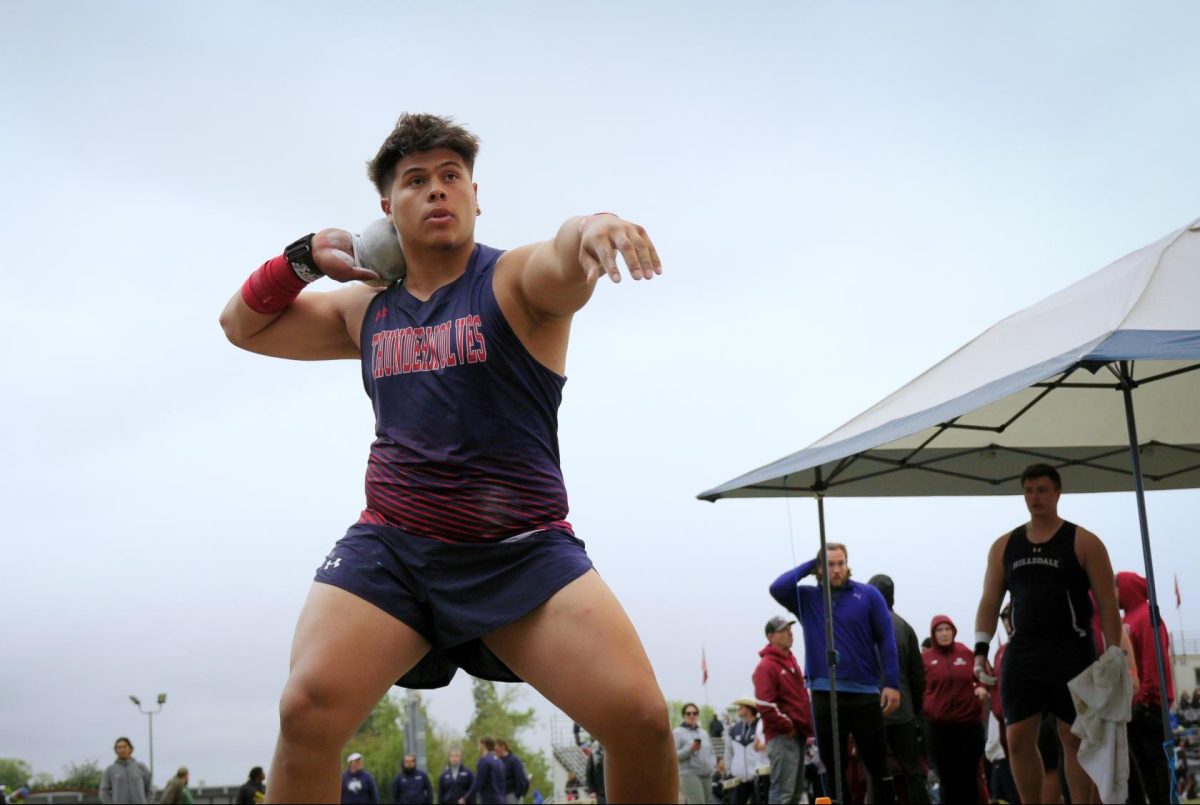 A man in a blue and red track uniform stands with a shot put.