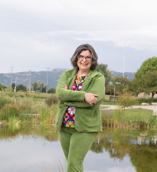 A woman stands in front of a small pond, wearing a green suit with a colorful shirt and smiling at the camera.