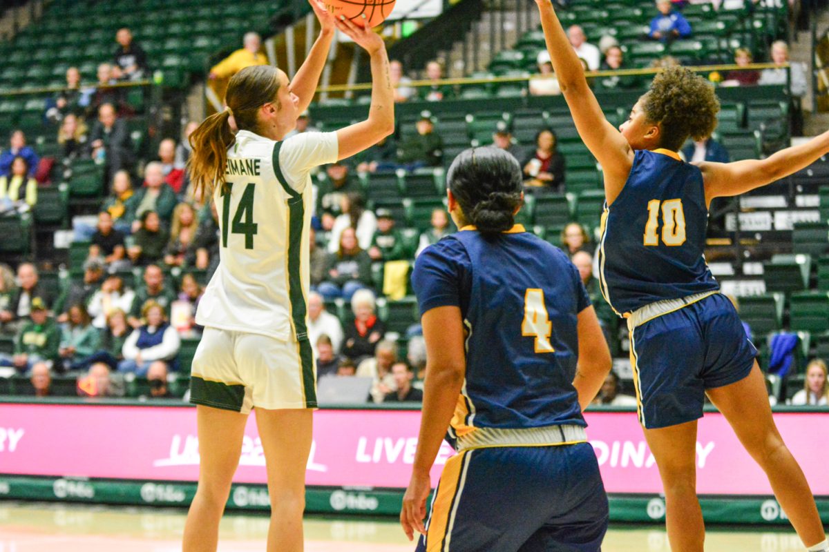 A girl in a green and white jersey shooting the ball while a girl in a blue and gold jersey tries to block her.