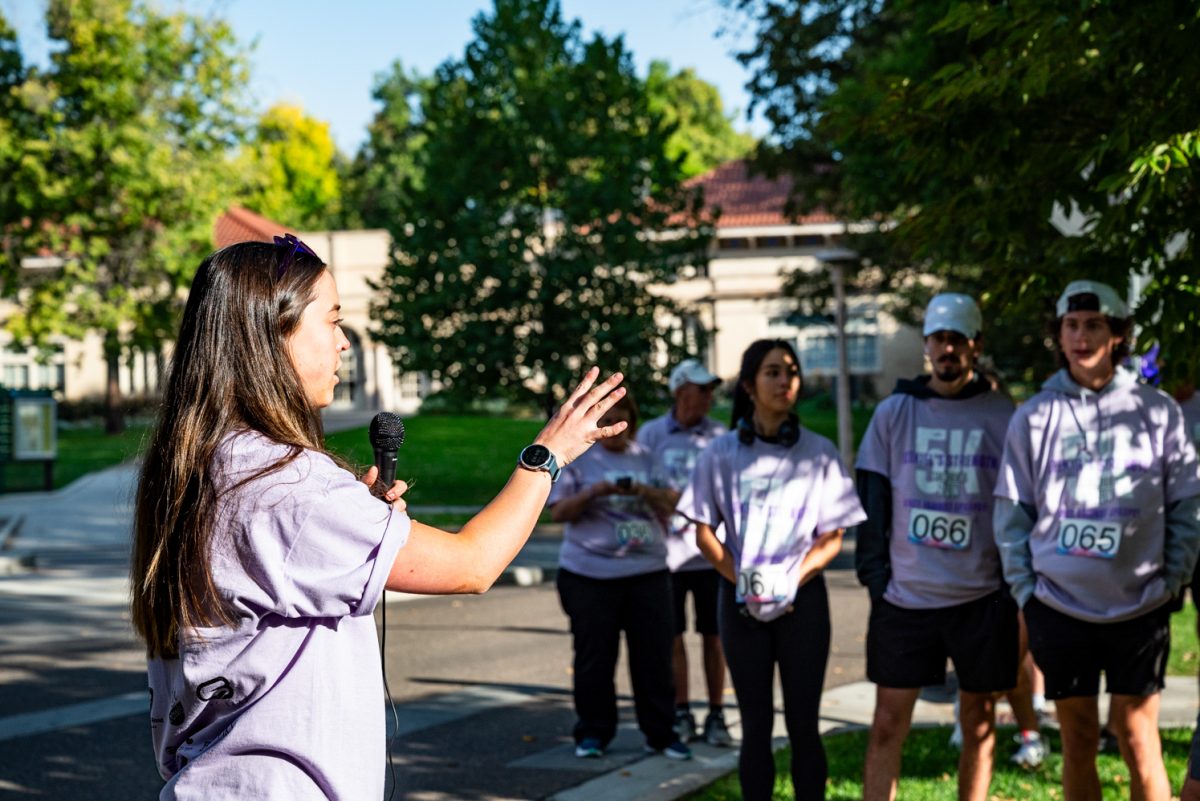 A woman in a light purple shirt speaks into a microphone and gestures to the handful of people gathered in front of her.