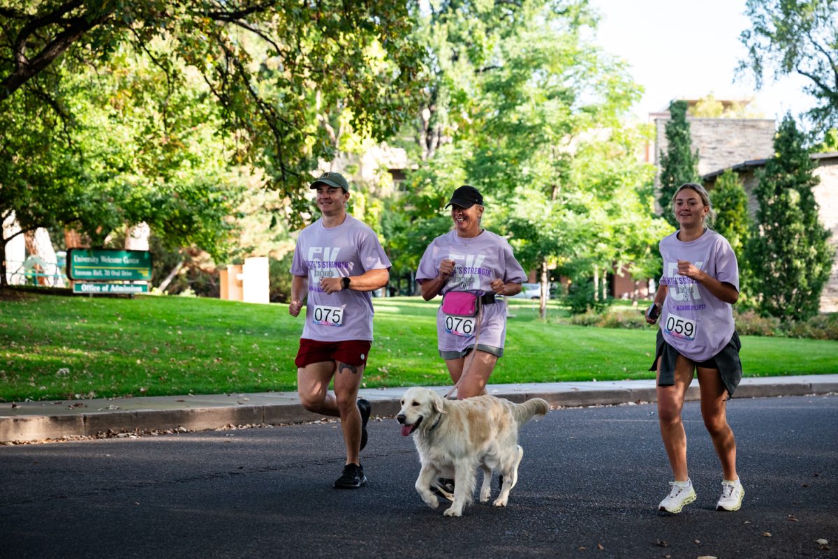 A man and two women in light purple shirts run together on a road. Their dog runs in front of them.