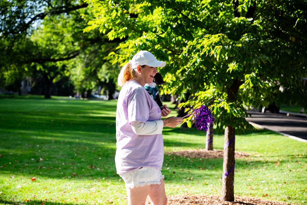 A woman in a light purple shirt and a white baseball cap holding a small black dog in a sweater and a purple pom pom.