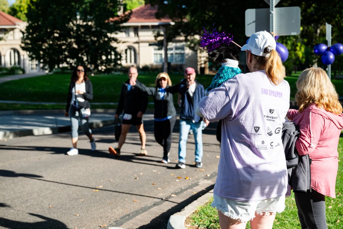 A woman in a light purple shirt waving a pom pom at a group of people walking by. She holds a small black dog in a sweater.