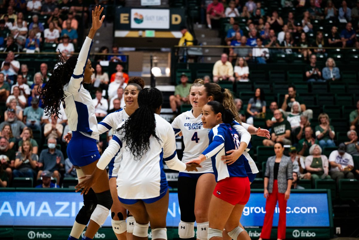 Volleyball players in white and blue huddle together and cheer. One player is jumping with her hands in the air.