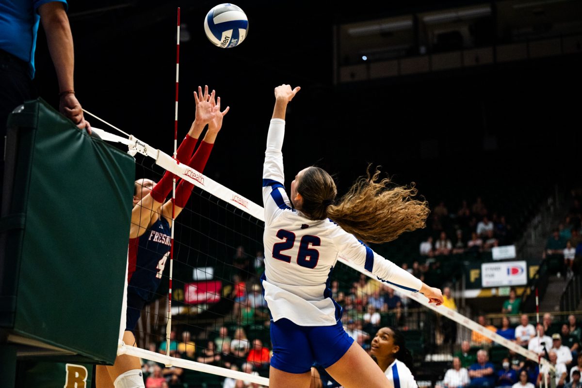 A volleyball player in white and blue jumps up above the net to hit a volleyball. On the other side of the net, a player in black and red jumps up as well.