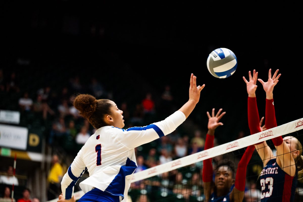 A volleyball player in white and blue jumps above the net and lightly hits a volleyball. On the other side of the net, two players in black and red jump up to block it.