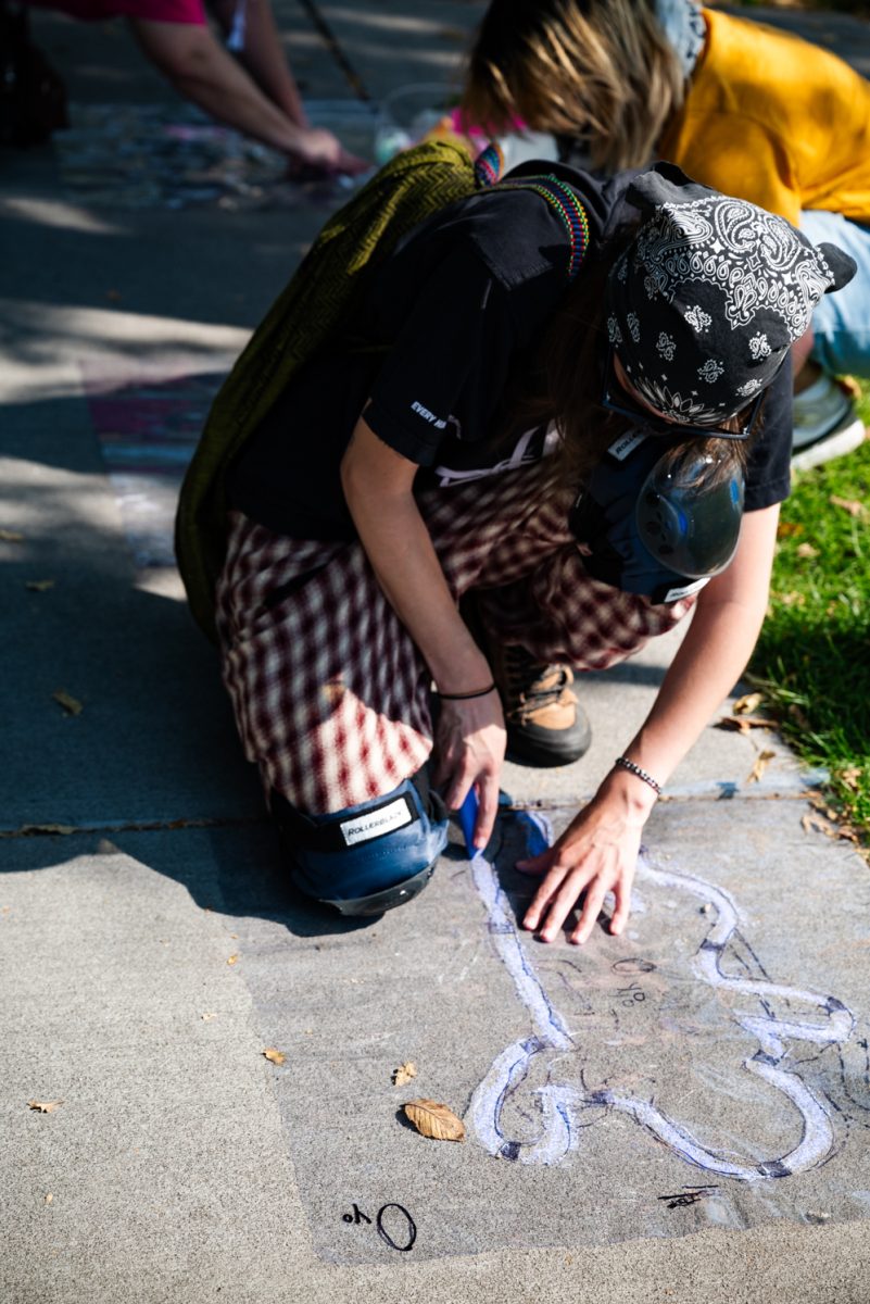 A person kneels on the sidewalk tracing an outline in purple chalk.