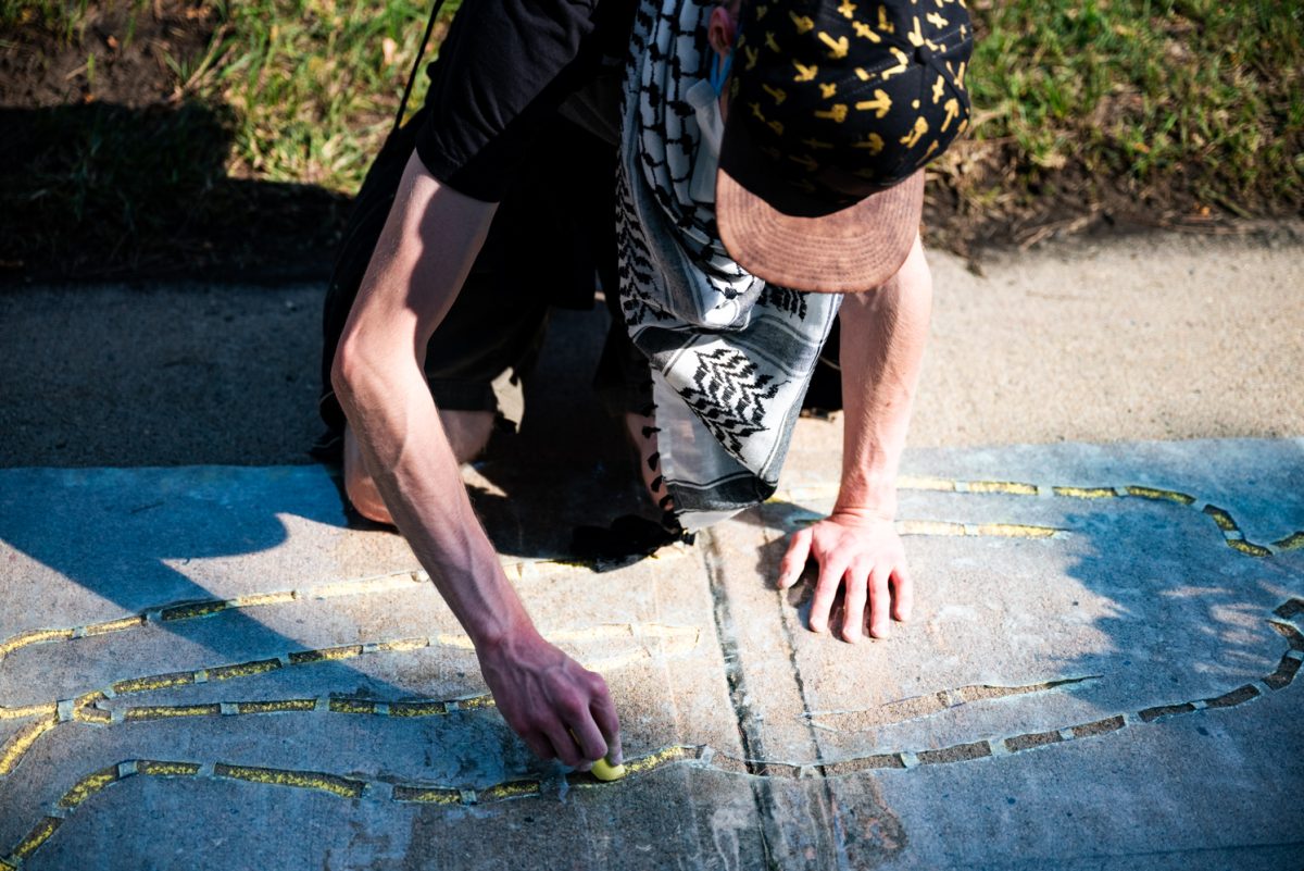 A person wearing a hat and a black and white scarf on their hands and knees drawing an outline of a body in yellow chalk.