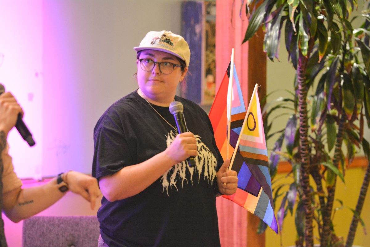 A speaker holding a microphone looking at her costar while holding the queer flags.
