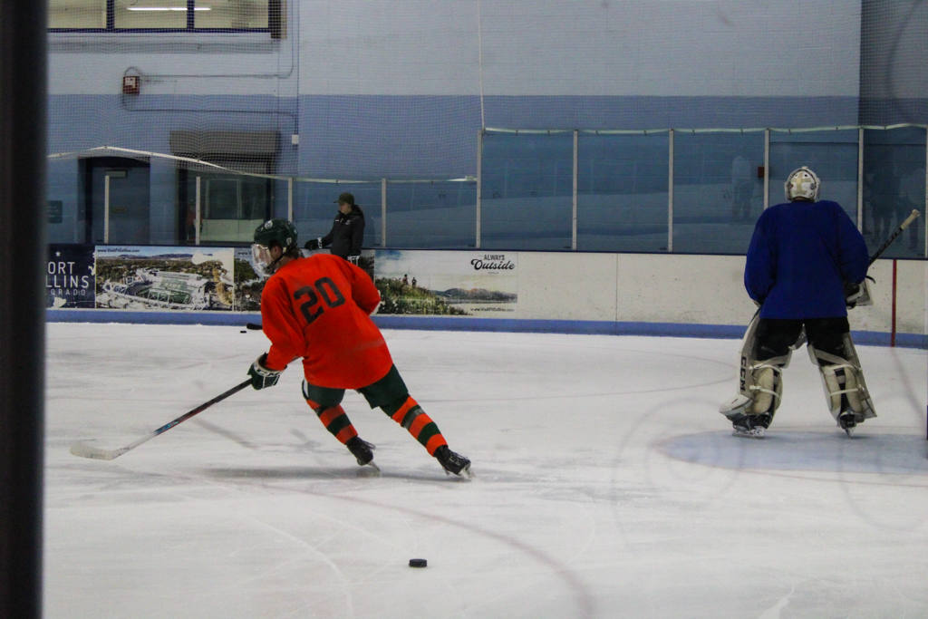 Two hockey players, one wearing orange and the other wearing blue, skate on an ice rink