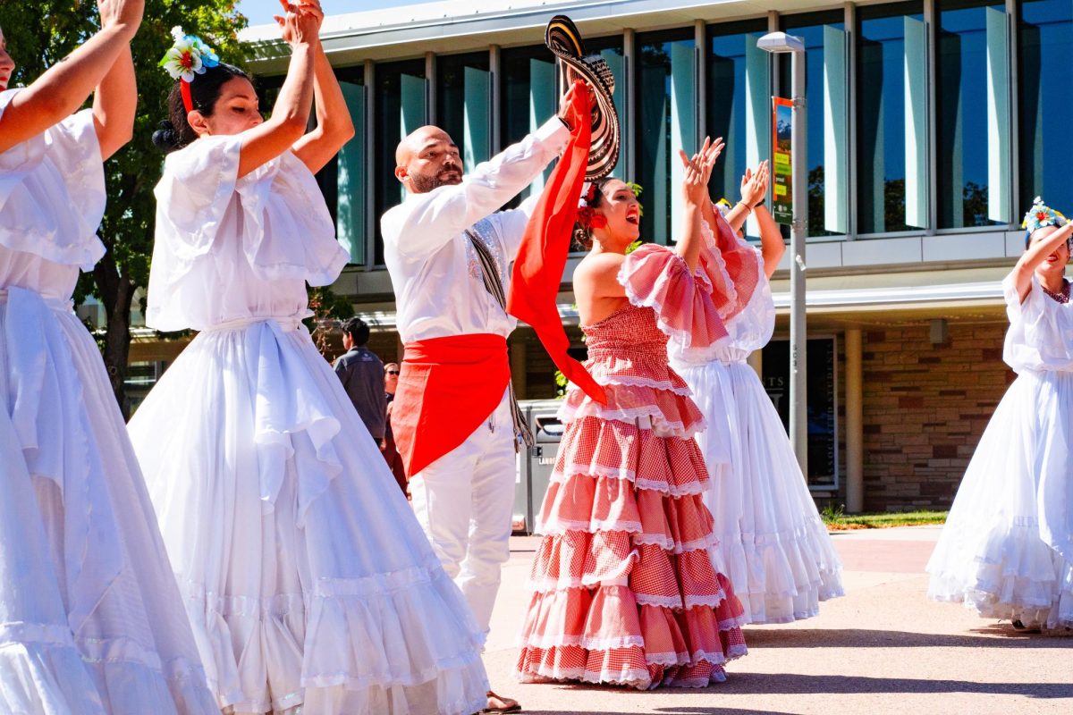 Several women in white dresses with big flowy, ruffled skirts dance with a man wearing a white shirt and pants with a red cloth around his wait, plus another woman in a red ruffled dress.