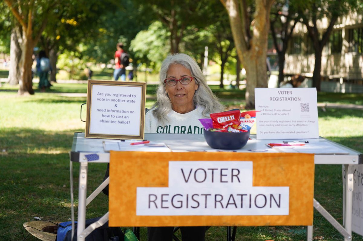 A woman with gray hair and red glasses sits at a table outside. The table displays signs reading, "Voter Registration", and "Are you registered to vote in another state & need information on how to cast an absentee ballot?".