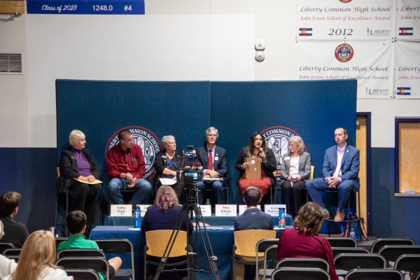 group of individuals sit on a stage with blue background and table with names in front of them, crowd in foreground