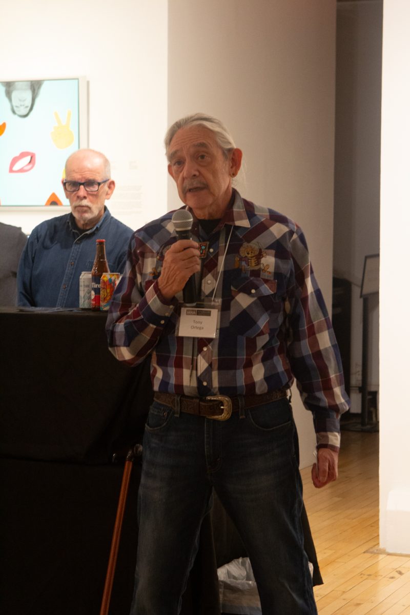 An older man holds a microphone and speaks while others stand beside and behind him, listening.