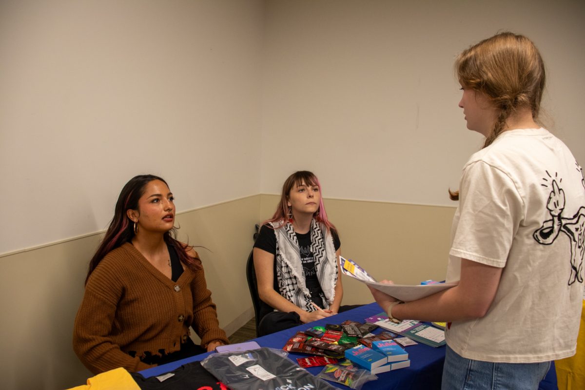 Two people sitting behind a table with giveaway items on it while a person in a white T-shirt stands holding a sheet of paper, talking.
