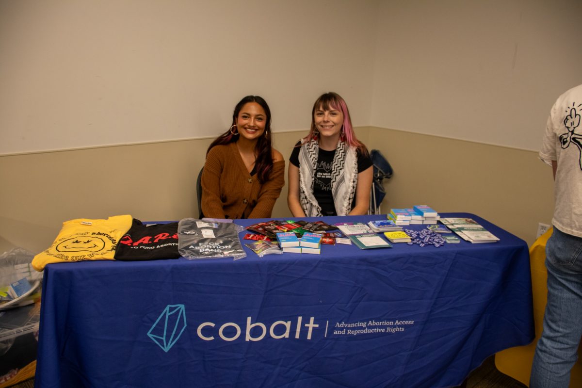 Two people sitting behind a table with things on it.