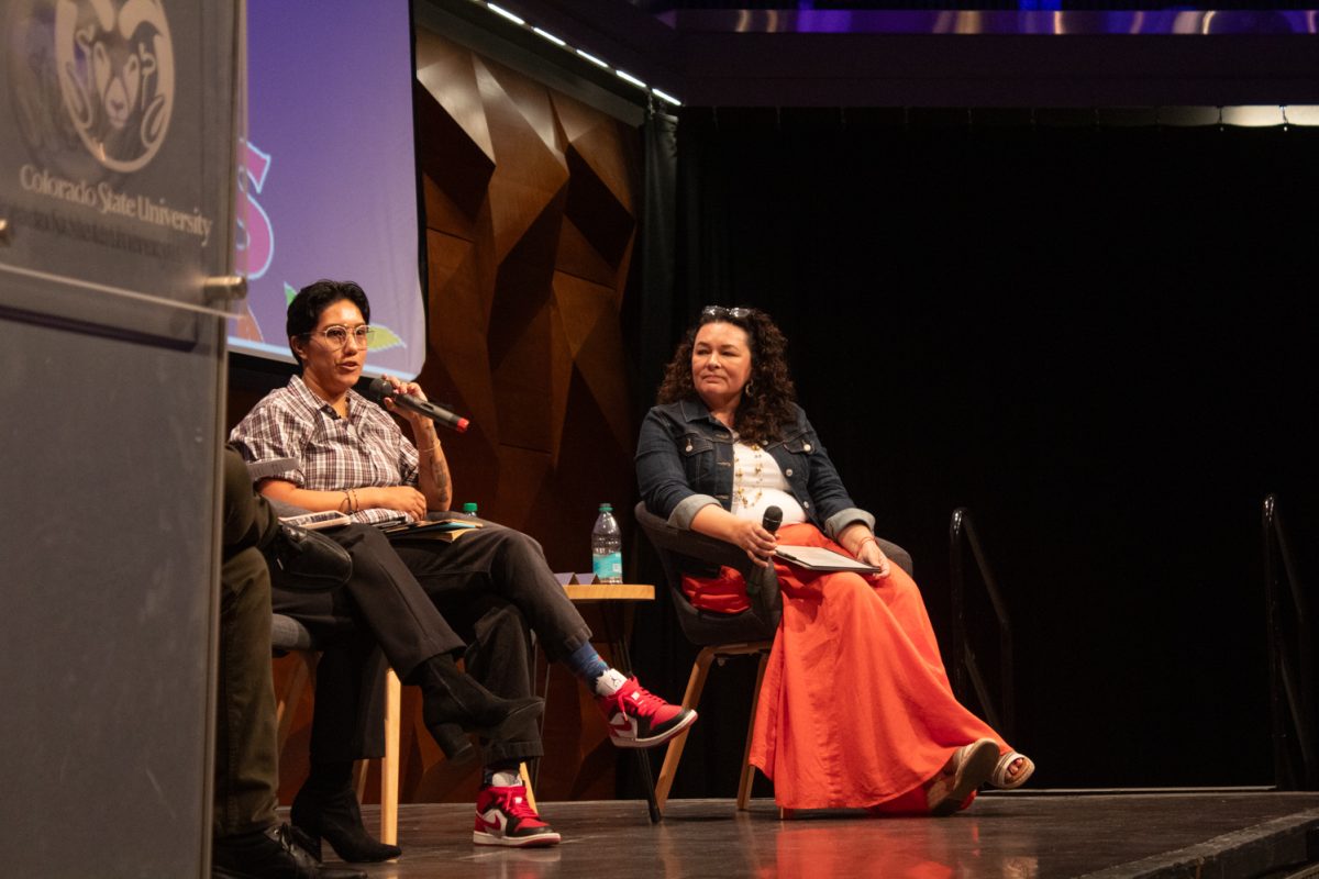 A gray podium with a piece of square glass displaying the CSU logo in white letters stands on a stage. A person with short black hair, glasses, a brown plaid button down shirt, brown pants and red sneakers sits in a chair on a stage holding a microphone to their mouth to the right of the podium. A woman with brown curly hair sits to the right of them wearing a blue denim jacket, a white shirt and a long orange skirt while also holding a microphone.