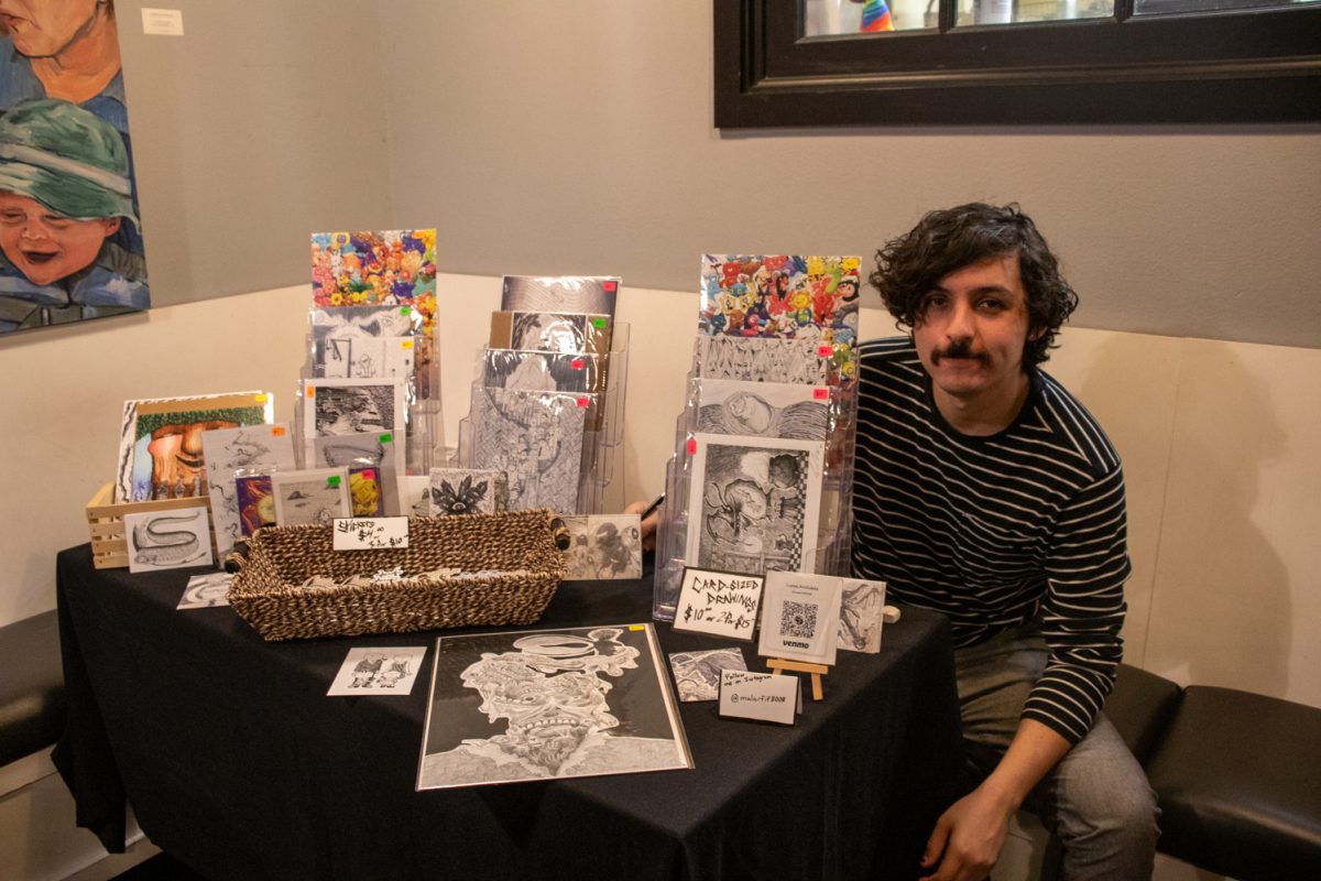 A person sitting behind a table with prints, images, and stickers for sale.
