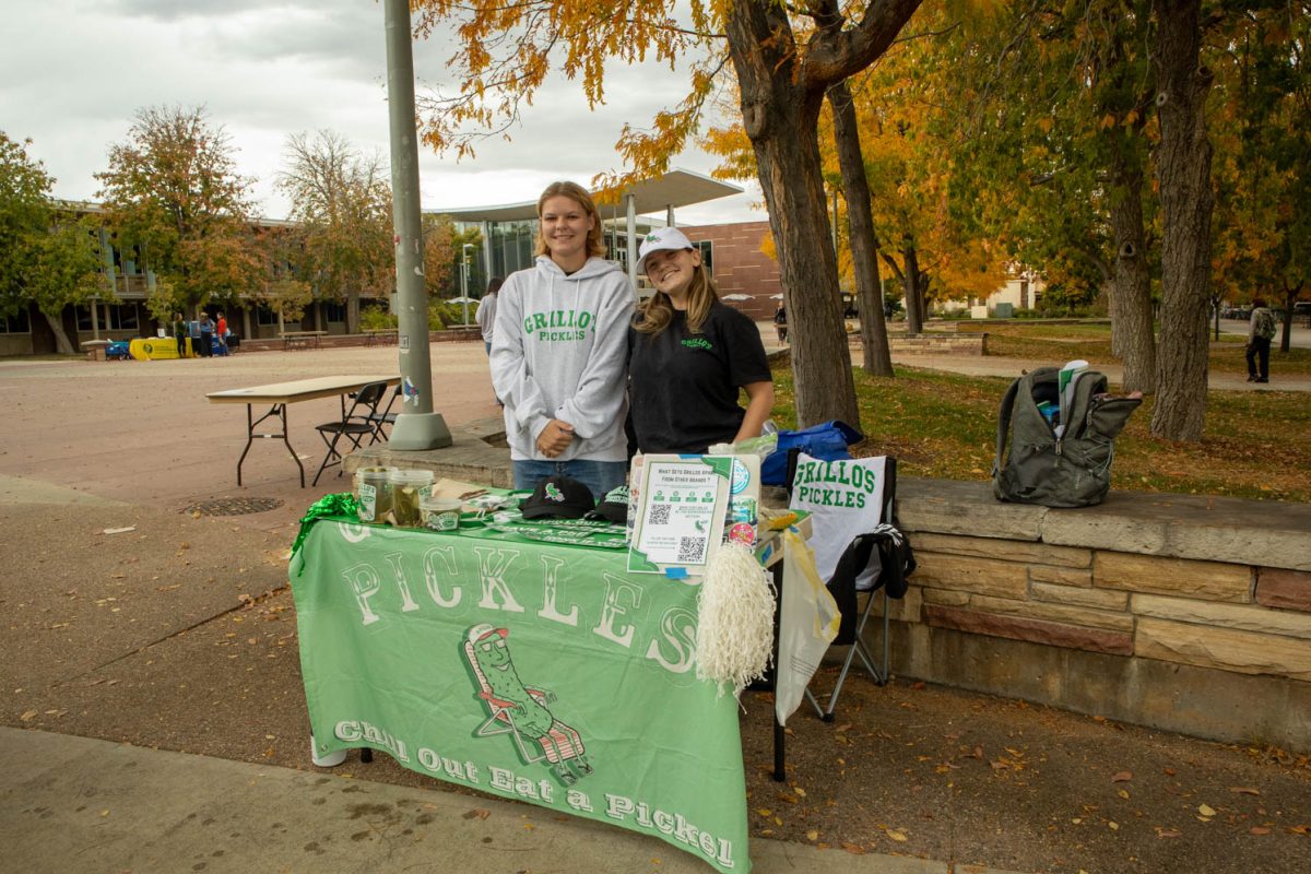 Two students stand behind a promotional table with a green cloth over the top that reads "Pickles" and "Chill Out Eat a Pickle."