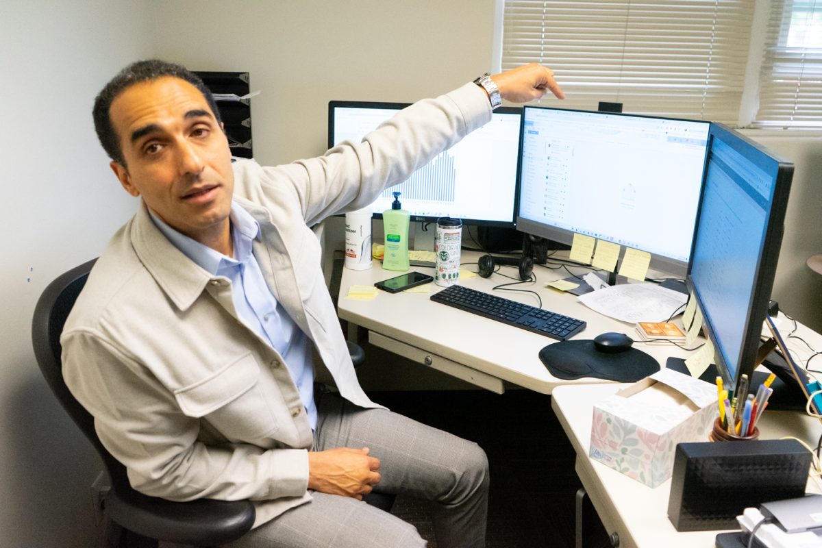 Man wearing a grey suit sitting at a desk, points at his computer.