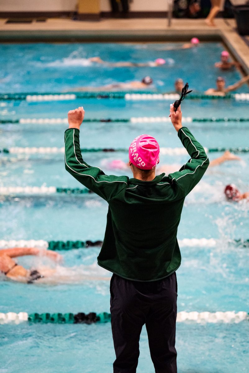 A swimmer in a green jacket holding her goggles cheers for her teammates swimming in the pool in front of her.