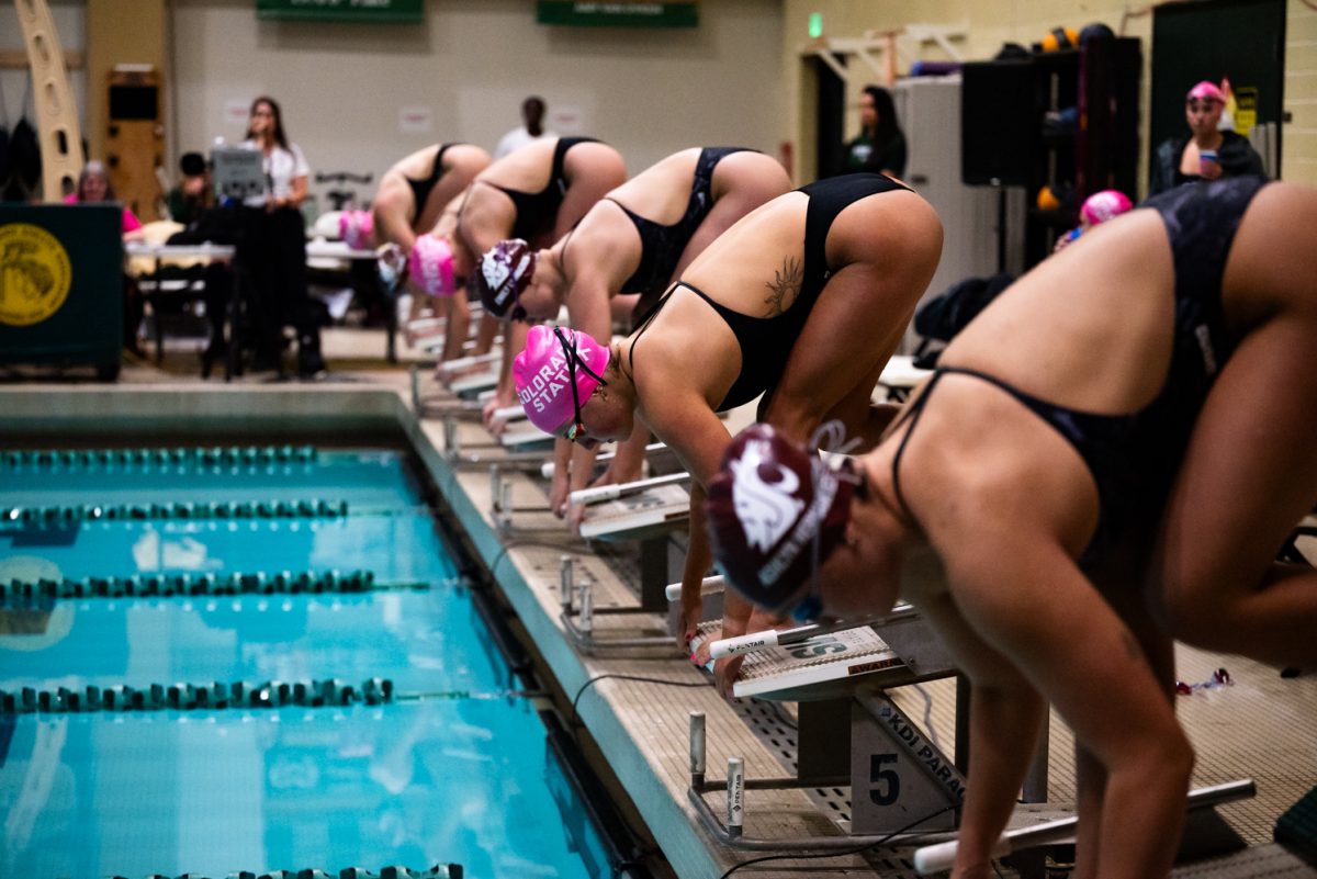 A row of five swimmers crouch on swim blocks getting ready to dive into a pool.