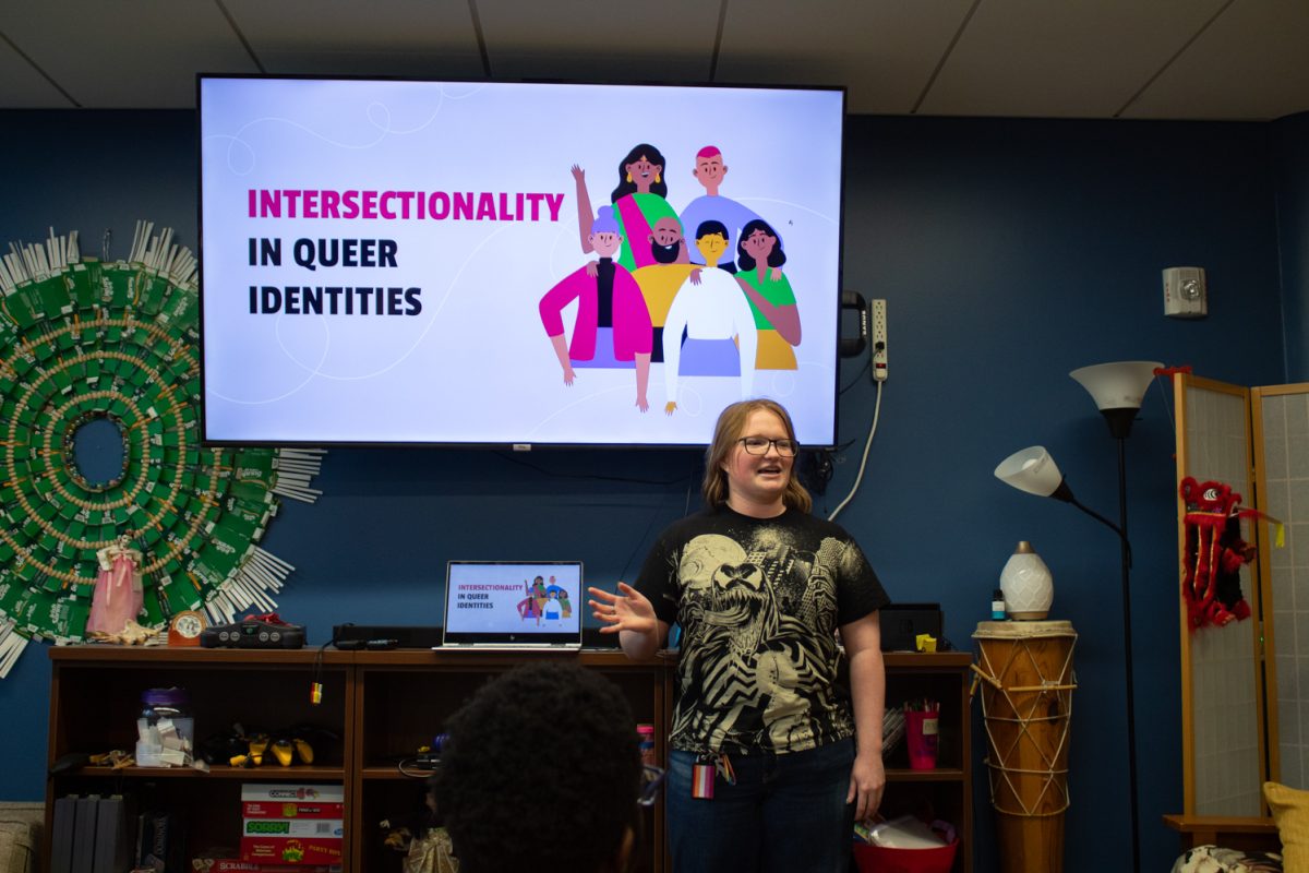 A person wearing a black t-shirt and jeans stands in front of a TV screen with a graphic of a group of people and pink and blue words. The words read, "Intersectionality in Queer Identities".