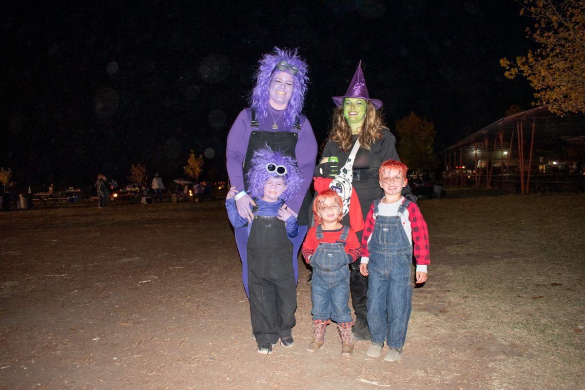 Two women stand with their sons. All of them are in Halloween costumes, and they are standing in an outdoor field area.