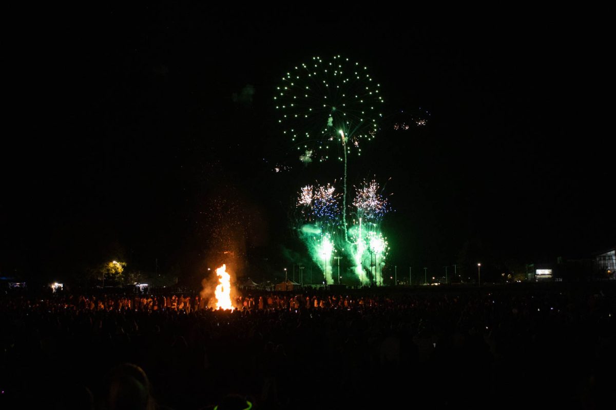 A large group of people surround a large fire as fireworks explode in the distance