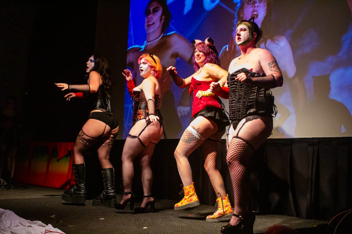 Four people wearing red and black outfits dance on stage while a movie is projected behind them in a dark theater.